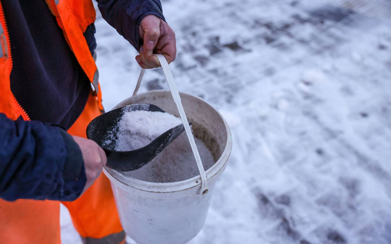Ein Mitarbeiter des Winterdienstes verteilt Streusalz bei Regen und Schneeglätte auf den Fußweg an einer Straßenkreuzung.