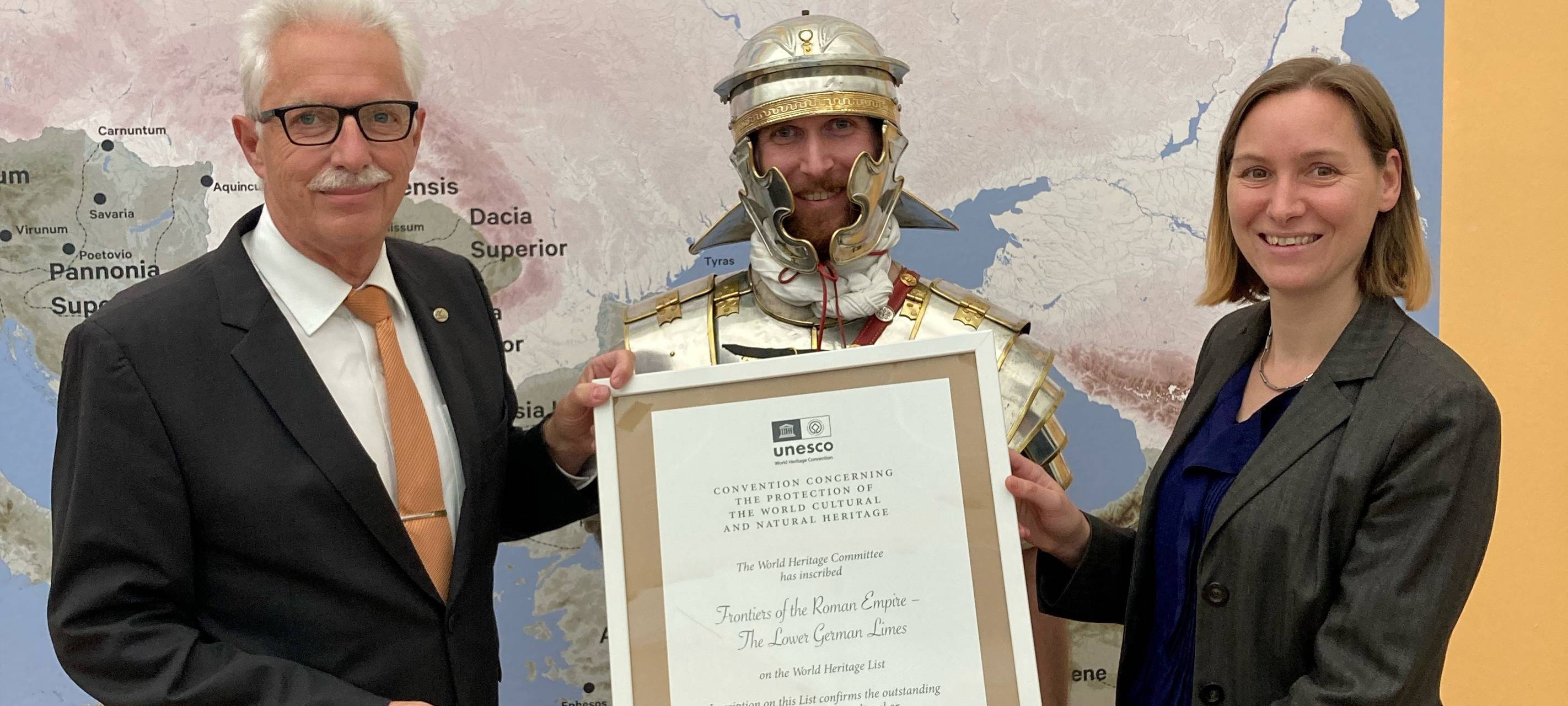 Landrat Thomas Hendele und Estelle Dageroth, Wirtschaftsförderung der Stadt Monheim am Rhein mit römischer Unterstützung halten die offizielle Urkunde in Händen.