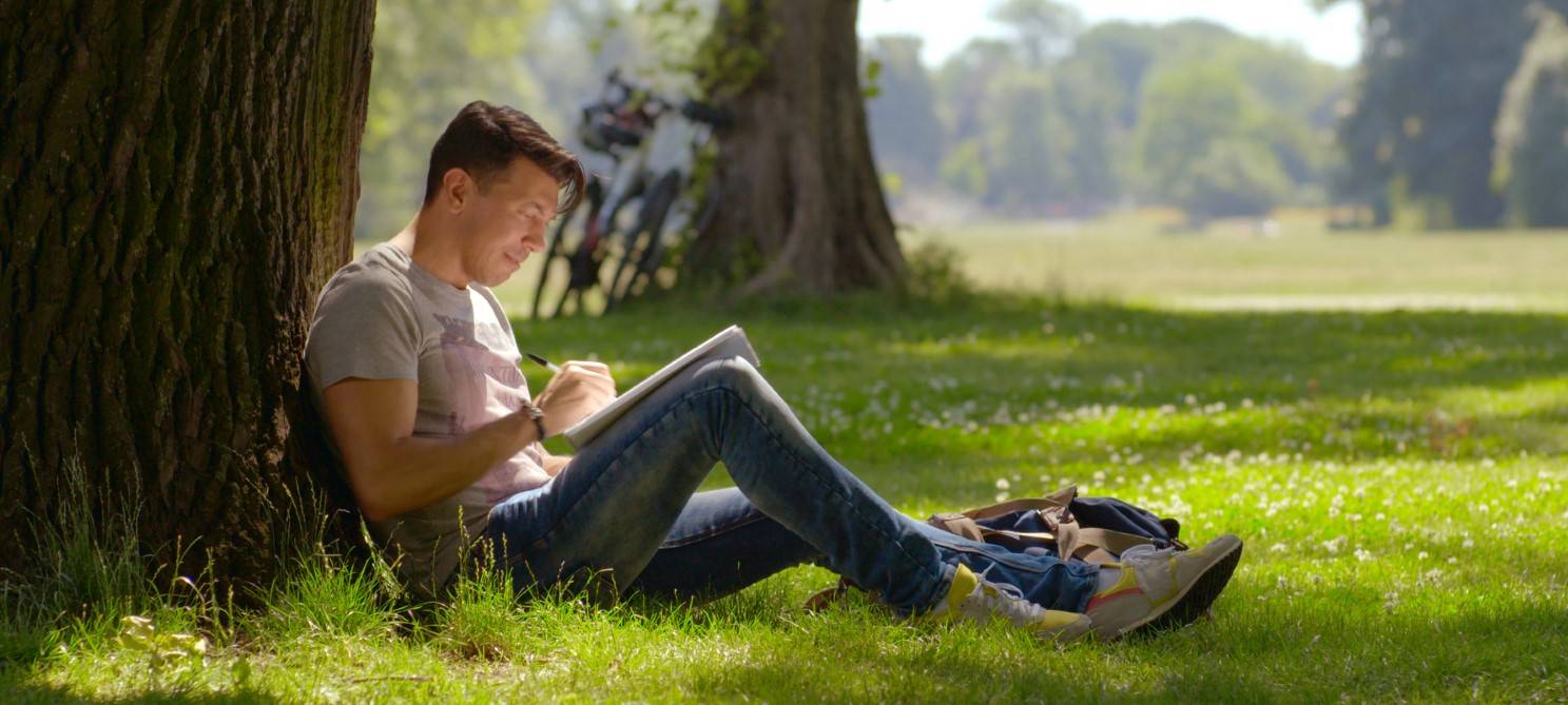 Alper sitzt an einem Baum auf einer Wiese und büffelt seinen Text
