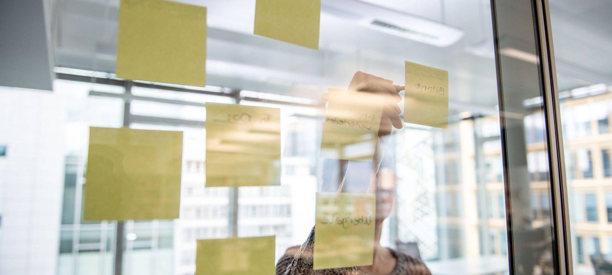 Eine Person klebt einen gelben Notizzettel an eine Glaswand